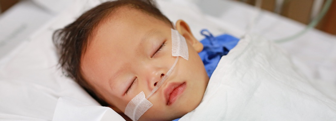 Close-up of a toddler with his eyes closed, laying in a hospital bed with an oxygen tube taped up to his nose.